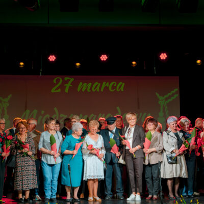 Szerokie ujęcie grupy kilkunastu osób stojących ramię w ramię na oświetlonej scenie. Nad grupą, na ekranie w tle, widnieje zielony napis „27 marca Międzynarodowy Dzień Teatru". Uczestnicy są uśmiechnięci, ubrani w różnorodne, eleganckie stroje, od garniturów po sukienki. Scena jest oświetlona ciepłym światłem scenicznym z różowymi akcentami.