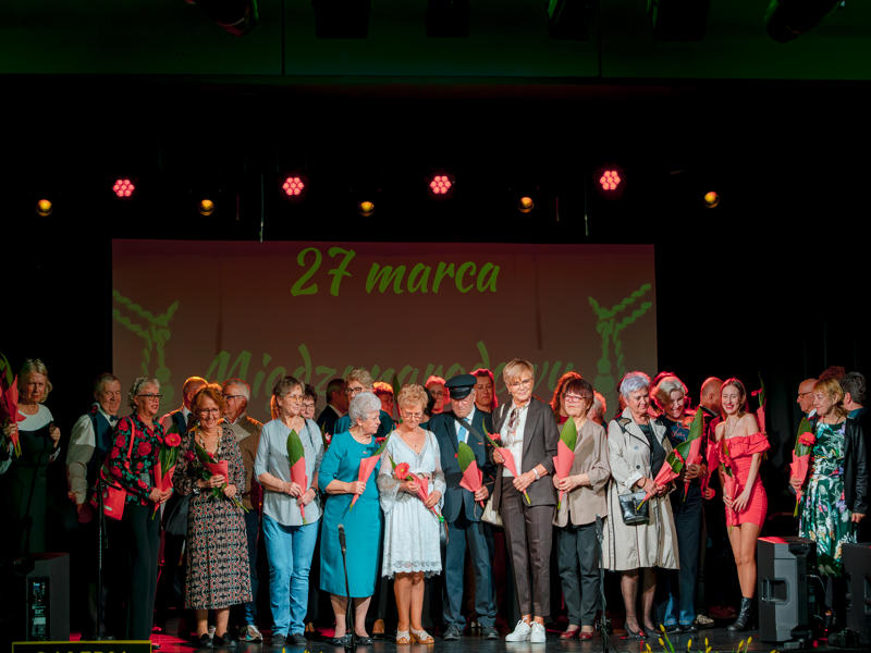 Szerokie ujęcie grupy kilkunastu osób stojących ramię w ramię na oświetlonej scenie. Nad grupą, na ekranie w tle, widnieje zielony napis „27 marca Międzynarodowy Dzień Teatru". Uczestnicy są uśmiechnięci, ubrani w różnorodne, eleganckie stroje, od garniturów po sukienki. Scena jest oświetlona ciepłym światłem scenicznym z różowymi akcentami.