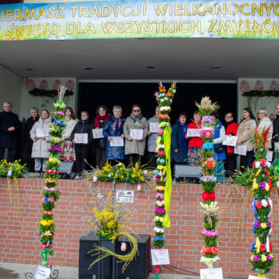 Szerokie ujęcie przedstawiające grupę kilkunastu osób (głównie kobiet) stojących w rzędzie na na scenie amfiteatru. Prawie każda osoba trzyma w dłoniach biały dyplom. Nad grupą rozpięty jest duży, żółto-biały baner z napisem: „KIERMASZ TRADYCJI WIELKANOCNYCH I ŚWIĘTO DLA WSZYSTKICH ZMYSŁÓW". Na pierwszym planie, przed sceną, ustawiono szereg bardzo wysokich, bogato zdobionych palm wielkanocnych, sięgających niemal dachu. Palmy są kolorowe, udekorowane sztucznymi kwiatami, gałązkami i wstążkami. Po prawej stronie wyróżnia się osoba w całkowicie zielonym stroju, z kapeluszem gęsto pokrytym różowymi kwiatami, trzymająca mikrofon.