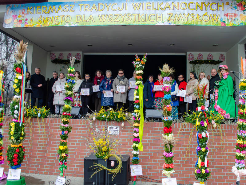 Szerokie ujęcie przedstawiające grupę kilkunastu osób (głównie kobiet) stojących w rzędzie na na scenie amfiteatru. Prawie każda osoba trzyma w dłoniach biały dyplom. Nad grupą rozpięty jest duży, żółto-biały baner z napisem: „KIERMASZ TRADYCJI WIELKANOCNYCH I ŚWIĘTO DLA WSZYSTKICH ZMYSŁÓW". Na pierwszym planie, przed sceną, ustawiono szereg bardzo wysokich, bogato zdobionych palm wielkanocnych, sięgających niemal dachu. Palmy są kolorowe, udekorowane sztucznymi kwiatami, gałązkami i wstążkami. Po prawej stronie wyróżnia się osoba w całkowicie zielonym stroju, z kapeluszem gęsto pokrytym różowymi kwiatami, trzymająca mikrofon.
