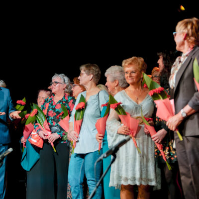 Wójt Gminy Grębocice Roman Jabłoński przemawia do grupy kobiet stojących na scenie. Panie są uśmiechnięte i trzymają pojedyncze czerwone kwiaty (gerbery) ozdobione zielonymi liśćmi i różowym papierem. Tło jest ciemne, typowe dla oprawy scenicznej.