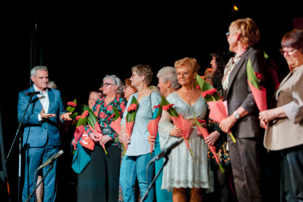 Wójt Gminy Grębocice Roman Jabłoński przemawia do grupy kobiet stojących na scenie. Panie są uśmiechnięte i trzymają pojedyncze czerwone kwiaty (gerbery) ozdobione zielonymi liśćmi i różowym papierem. Tło jest ciemne, typowe dla oprawy scenicznej.