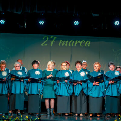 Chór mieszany występujący na scenie. Większość chórzystów to kobiety ubrane w identyczne błękitne stroje z białą ozdobą. Wszyscy trzymają otwarte teczki z nutami. W centralnym punkcie stoi kobieta, trzymająca mikrofon.