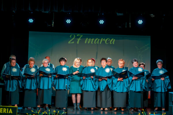 Chór mieszany występujący na scenie. Większość chórzystów to kobiety ubrane w identyczne błękitne stroje z białą ozdobą. Wszyscy trzymają otwarte teczki z nutami. W centralnym punkcie stoi kobieta, trzymająca mikrofon.