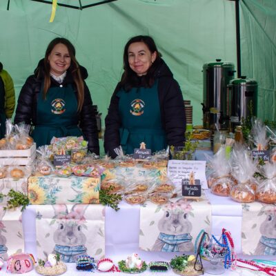 Trzy uśmiechnięte kobiety za ladą stoiska sprzedażowego. Ubrane są w jednakowe ciemnozielone fartuchy z logo. Na białym obrusie z motywem zajączków wyeksponowane są produkty: domowe wypieki w celofanowych woreczkach (babeczki, mazurki) oraz ręcznie robiona biżuteria i ozdoby z koralików. Widoczne są małe tabliczki z cenami.