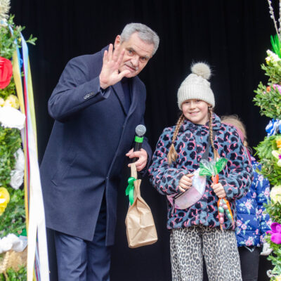 Wójt gminy, w ciemnym płaszczu uśmiecha się i macha ręką do obiektywu. W drugiej dłoni trzyma mikrofon i papierową torebkę. Obok niego stoi uśmiechnięta dziewczynka w jasnej czapce z pomponem i wzorzystej kurtce, trzymająca upominek zapakowany w celofan. Po lewej stronie widoczna jest wysoka, kolorowa palma wielkanocna.