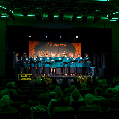 Szeroki plan na salę widowiskową. Na oświetlonej zielonymi neonami scenie występuje chór składający się z kilkunastu osób ubranych w błękitne stroje. W tle widnieje napis „27 marca Międzynarodowy Dzień Teatru". Widownia siedzi w fotelach, tyłem do obiektywu, obserwując występ.