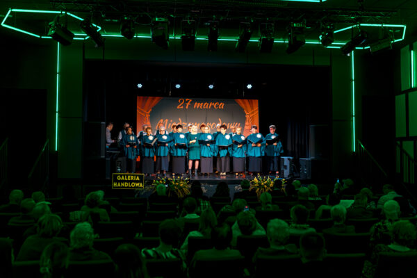 Szeroki plan na salę widowiskową. Na oświetlonej zielonymi neonami scenie występuje chór składający się z kilkunastu osób ubranych w błękitne stroje. W tle widnieje napis „27 marca Międzynarodowy Dzień Teatru". Widownia siedzi w fotelach, tyłem do obiektywu, obserwując występ.