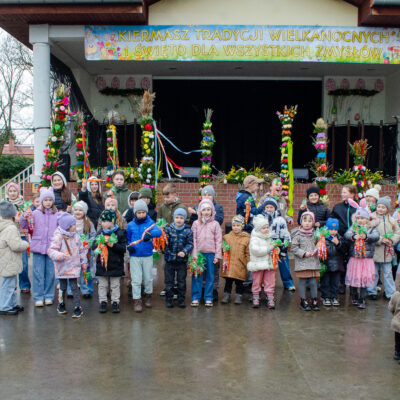 Szerokie ujęcie plenerowej sceny podczas „Kiermaszu Tradycji Wielkanocnych". Na pierwszym planie, przed ceglanym podestem, stoi duża grupa dzieci w różnym wieku, ubranych w kolorowe kurtki i czapki. Wiele z nich trzyma upominki w kształcie marchewek. Scena udekorowana jest licznymi, wysokimi palmami wielkanocnymi oraz napisem okolicznościowym na górze.