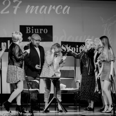 Czarno-białe zdjęcie ze spektaklu teatralnego. Grupa aktorów w różnym wieku znajduje się w dynamicznych pozach wokół stolika. Jedna z kobiet trzyma mikrofon, inna wykonuje ekspresyjne gesty rękami. W tle widoczne są tabliczki z napisami „Biuro", „Bank" oraz „Szpital".