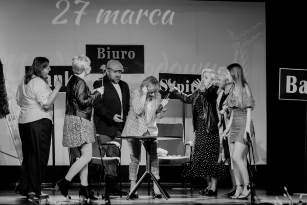 Czarno-białe zdjęcie ze spektaklu teatralnego. Grupa aktorów w różnym wieku znajduje się w dynamicznych pozach wokół stolika. Jedna z kobiet trzyma mikrofon, inna wykonuje ekspresyjne gesty rękami. W tle widoczne są tabliczki z napisami „Biuro", „Bank" oraz „Szpital".