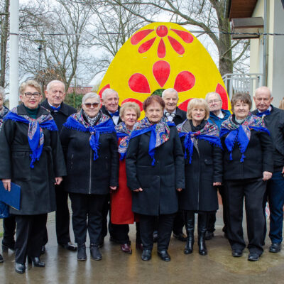 Pamiątkowe zdjęcie grupy seniorów (mężczyzn i kobiet) stojących na zewnątrz. Kobiety mają na ramionach jednakowe, niebieskie chusty z ludowymi wzorami, a mężczyźni pod szyjami niebieskie muchy. W tle widać dekorację w kształcie wielkiego, żółtego jaja wielkanocnego z czerwonymi zdobieniami.