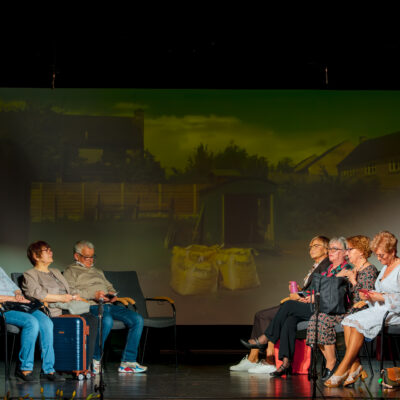 Scena ze spektaklu. Dwie grupy osób siedzą na krzesłach naprzeciwko siebie. Pośrodku, na ekranie w tle, wyświetlane jest zdjęcie wiejskiego podwórka z żółtymi workami. Aktorzy są ubrani w codzienne stroje, obok jednej z kobiet stoi niebieska walizka.
