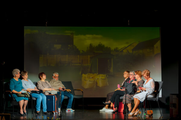 Scena ze spektaklu. Dwie grupy osób siedzą na krzesłach naprzeciwko siebie. Pośrodku, na ekranie w tle, wyświetlane jest zdjęcie wiejskiego podwórka z żółtymi workami. Aktorzy są ubrani w codzienne stroje, obok jednej z kobiet stoi niebieska walizka.