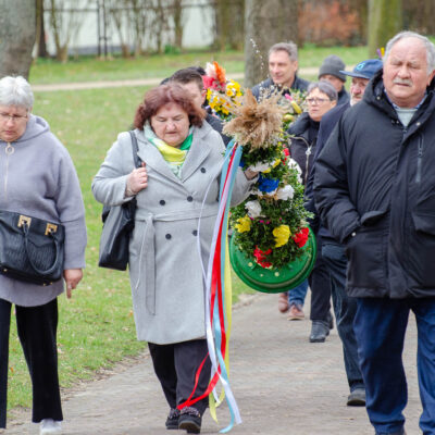Grupa osób idąca chodnikiem w parku lub ogrodzie. Na pierwszym planie kobieta w szarym płaszczu niesie dużą palmę wielkanocną ozdobioną kolorowymi wstążkami i kwiatami. Pozostali uczestnicy idą za nią w luźnym szyku. Pogoda wydaje się pochmurna, drzewa w tle są bez liści.
