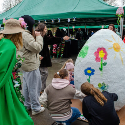 Grupa dzieci i dorosłych zgromadzona pod zielonym namiotem wokół ogromnej makiety białego jajka wielkanocnego. Uczestnicy malują na nim kolorowe kwiaty i słońce. Po lewej stronie tyłem stoi osoba w jaskrawozielonym stroju i kapeluszu ozdobionym kwiatami, obserwująca proces twórczy.