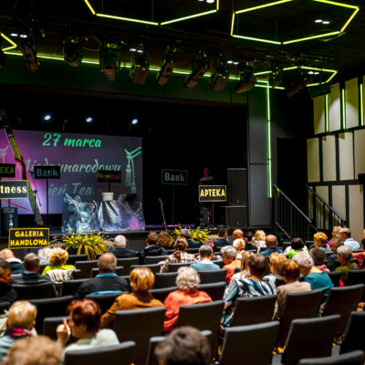 Widok z tyłu sali na wypełnioną widownię i scenę. Scena przygotowana jest do występu. Znajdują się na niej rekwizyty przykryte folią oraz tabliczki informacyjne (m.in. „Apteka", „Fitness", „Bank").