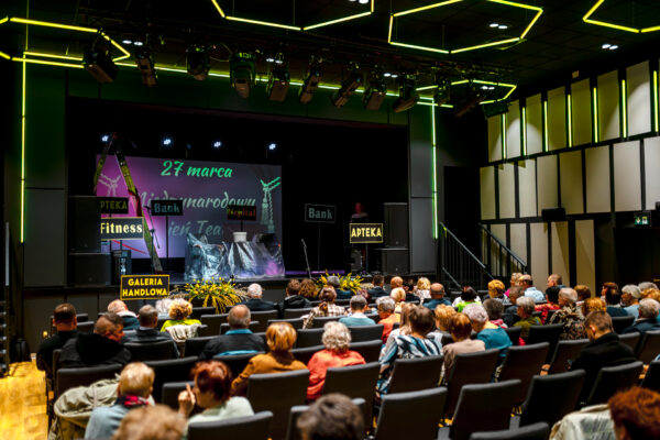 Widok z tyłu sali na wypełnioną widownię i scenę. Scena przygotowana jest do występu. Znajdują się na niej rekwizyty przykryte folią oraz tabliczki informacyjne (m.in. „Apteka", „Fitness", „Bank").