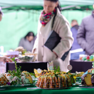 Zbliżenie na stół przykryty zielonym obrusem, na którym wyeksponowane są tradycyjne babki wielkanocne. Ciasta mają różne polewy i są udekorowane zielonymi gałązkami oraz ozdobami na patyczkach. W tle, w rozmyciu, widać osoby odwiedzające stoiska kiermaszowe pod namiotami.