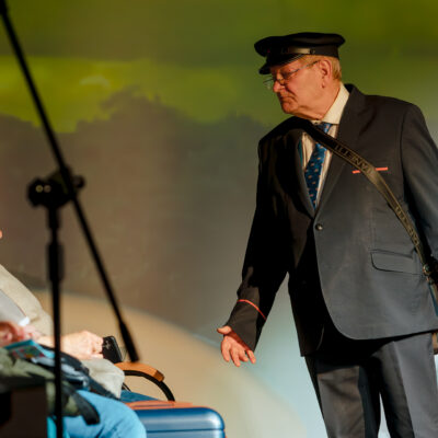 Zbliżenie na aktora grającego rolę konduktora. Mężczyzna ma na sobie ciemny mundur, czapkę z daszkiem i torbę przewieszoną przez ramię. Stoi przed siedzącymi na scenie pasażerami, wykonując zapraszający gest dłonią.