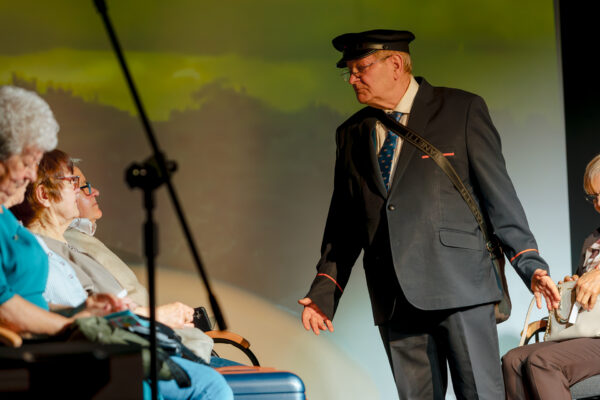 Zbliżenie na aktora grającego rolę konduktora. Mężczyzna ma na sobie ciemny mundur, czapkę z daszkiem i torbę przewieszoną przez ramię. Stoi przed siedzącymi na scenie pasażerami, wykonując zapraszający gest dłonią.