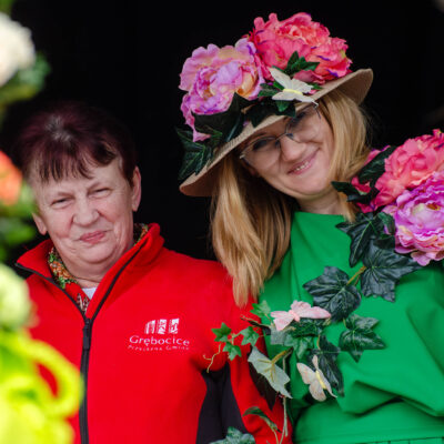 Portret dwóch uśmiechniętych kobiet. Kobieta po prawej ma na sobie jaskrawozielony strój oraz słomkowy kapelusz gęsto zdobiony dużymi, różowymi kwiatami. Kobieta po lewej ubrana jest w czerwoną bluzę z logo „Grębocice". Wokół nich widać fragmenty kolorowych dekoracji kwiatowych.