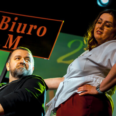 Portret dwójki aktorów na scenie. Widok z dołu. Mężczyzna z brodą patrzy w górę, a stojąca obok niego kobieta w okularach i jasnej koszuli trzyma się pod boki, patrząc na niego z góry. W tle widoczny fragment napisu „Biuro M.".