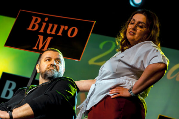 Portret dwójki aktorów na scenie. Widok z dołu. Mężczyzna z brodą patrzy w górę, a stojąca obok niego kobieta w okularach i jasnej koszuli trzyma się pod boki, patrząc na niego z góry. W tle widoczny fragment napisu „Biuro M.".