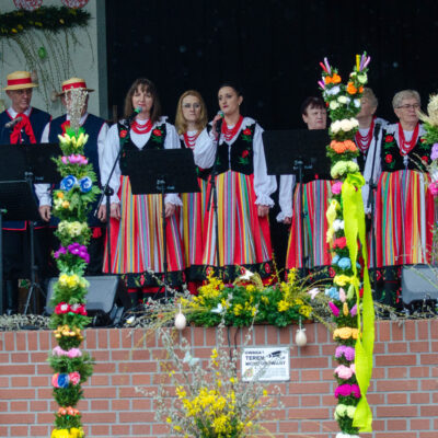Występ zespołu ludowego na scenie ozdobionej palmami i kwiatami. Mężczyźni ubrani są w ciemne kamizelki, białe koszule, czerwone chusty i słomkowe kapelusze. Kobiety mają na sobie tradycyjne stroje z haftowanymi gorsetami i kolorowymi spódnicami w pasy. Niektóre osoby śpiewają do mikrofonów.