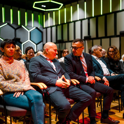 Widzowie siedzący w pierwszym rzędzie sali widowiskowej. W centralnej części widać dwóch mężczyzn w garniturach prowadzących rozmowę. Obok nich siedzi kobieta w kolorowym swetrze. W dalszych fotelach mężczyzna (Wójt Gminy) rozmawia z kobietą dyrektorką Centrum Kultury) Ściany sali ozdobione są panelami w kształcie plastra miodu i pionowymi pasami światła.