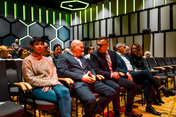 Widzowie siedzący w pierwszym rzędzie sali widowiskowej. W centralnej części widać dwóch mężczyzn w garniturach prowadzących rozmowę. Obok nich siedzi kobieta w kolorowym swetrze. W dalszych fotelach mężczyzna (Wójt Gminy) rozmawia z kobietą dyrektorką Centrum Kultury) Ściany sali ozdobione są panelami w kształcie plastra miodu i pionowymi pasami światła.