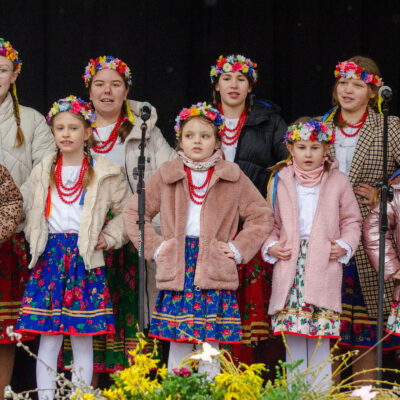 Grupa dziewczynek występująca na scenie w strojach ludowych. Mają na głowach kolorowe wianki z kwiatów, białe bluzki z czerwonymi koralami oraz wzorzyste spódniczki. Na wierzch założyły codzienne kurtki i płaszcze. Dziewczynki śpiewają, trzymając ręce na biodrach.