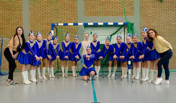 Zdjęcie grupowe zespołu mażoretek w hali sportowej. Piętnaście dziewcząt w niebiesko-złotych strojach z białymi wysokimi butami stoi w rzędzie, trzymając przed sobą pałeczki. Jedna z nich klęczy w centrum grupy. Po bokach stoją dwie opiekunki w strojach cywilnych. W tle widać bramkę do piłki ręcznej i ceglaną ścianę.