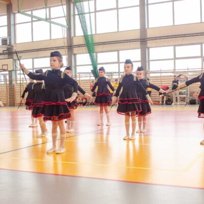 Grupa młodych mażoretek w ciemnogranatowych strojach z czerwonymi akcentami i furażerkach wykonuje układ taneczny na boisku hali sportowej. Dziewczynki są rozproszone po parkiecie, każda trzyma pałeczkę. W tle widać duże okna hali.