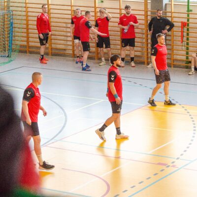 Widok z góry na grupę mężczyzn w czerwonych i czarnych koszulkach sportowych znajdujących się na boisku. Niektórzy zawodnicy rozmawiają, inni odpoczywają przy drabinkach gimnastycznych. Zdjęcie wykonane zza fragmentu nieostrej przeszkody na pierwszym planie.