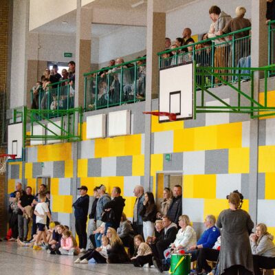 Wnętrze hali sportowej wypełnione ludźmi. Na parterze pod ścianą z żółto-szarych paneli oraz na balkonie z zieloną barierką stoją liczni widzowie obserwujący wydarzenie.