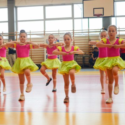 Grupa najmłodszych mażoretek w jaskrawych, różowo-żółtych strojach. Dziewczynki stoją na jednej nodze, trzymając pałeczki poziomo przed klatką piersiową, wykonując element choreografii.