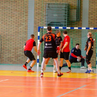 Dynamiczne ujęcie z meczu piłki ręcznej. Kilku zawodników w czerwonych i czarnych strojach znajduje się w pobliżu bramki. Bramkarz w niebieskiej bluzie jest w pozycji gotowej do obrony strzału.