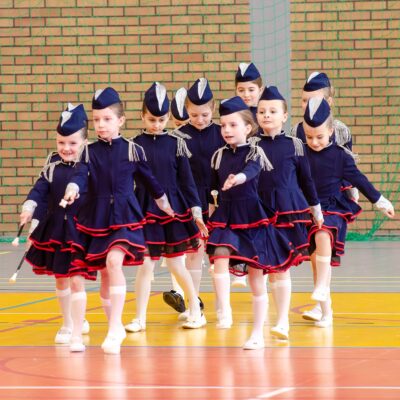 Zbliżenie na grupę mażoretek w granatowych mundurkach maszerujących w szyku. Dziewczynki mają skupione miny i wykonują zsynchronizowany krok.