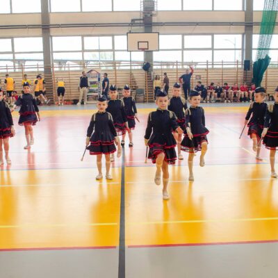 Mażoretki w granatowych strojach podczas występu na środku hali. Formacja przypomina trójkąt, a dziewczynki mają uniesione nogi w marszu. W tle widać sędziów lub trenerów przy stoliku oraz pozostałych uczestników pod oknami.