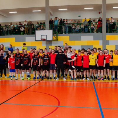 Duże zdjęcie grupowe wszystkich uczestników wydarzenia sportowego na boisku. W pierwszym rzędzie stoją zawodnicy w strojach sportowych (czerwonych, czarnych i żółtych), niektórzy trzymają dzieci na rękach. Za nimi, na trybunach i balkonach, widoczny jest tłum kibiców. Wszyscy patrzą w stronę aparatu.