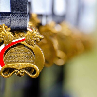 Zbliżenie na pamiątkowy złoty medal zawodów "Wolf Race – Szlakiem Dziadoszan". Medal ma kształt okręgu zwieńczonego dwiema głowami wilków, a przez środek przechodzi biało-czerwona wstęga. W tle widać rzędy kolejnych, rozmytych medali.