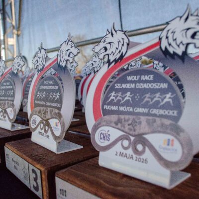 Rzędy białych statuetek z logotypem biegu "Wolf Race" i sylwetką biegnącego wilka, ustawione na drewnianych podstawach. Na statuetkach widnieje napis "Puchar Wójta Gminy Grębocice" oraz data "2 maja 2026".