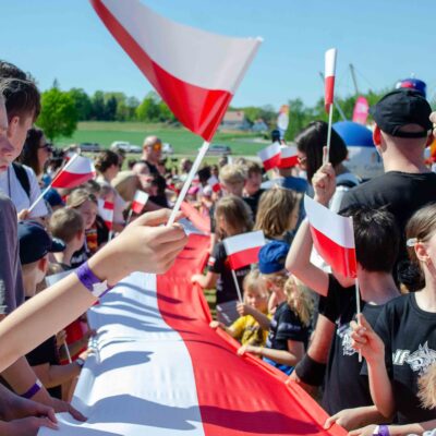 Tłum dzieci i dorosłych trzyma wspólnie ogromną, długą flagę Polski podczas wydarzenia plenerowego. Wiele osób w tłumie trzyma w dłoniach małe flagi narodowe na patyczkach.