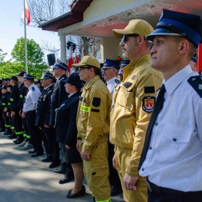 Szereg funkcjonariuszy różnych służb mundurowych stoi na baczność podczas uroczystości plenerowej. Na pierwszym planie mężczyzna w białej koszuli z krótkim rękawem, dalej strażacy w piaskowych mundurach bojowych oraz policjanci w granatowych mundurach wyjściowych. W tle widoczne poczty sztandarowe.