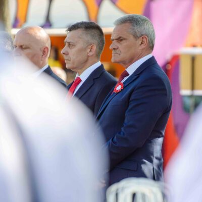 Trzej mężczyźni w ciemnych garniturach stoją z powagą w profilu podczas oficjalnego wydarzenia. Jeden z nich ma wpięty w klapę biało-czerwony kotylion. Pierwszy plan jest lekko rozmazany, co skupia uwagę na twarzach mężczyzn.