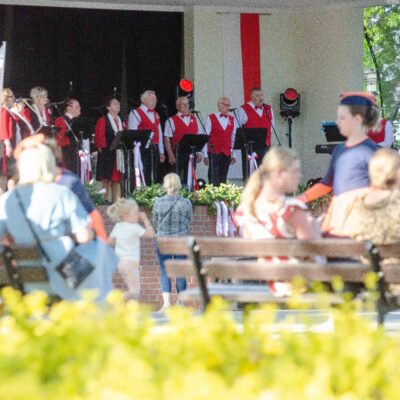 Widok zza pleców publiczności siedzącej na ławkach na scenę amfiteatru. Na scenie występuje zespół wokalny (mężczyźni i kobiety) ubrany w białe koszule i czerwone kamizelki. Scena udekorowana jest flagami Polski.