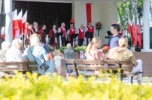 Widok zza pleców publiczności siedzącej na ławkach na scenę amfiteatru. Na scenie występuje zespół wokalny (mężczyźni i kobiety) ubrany w białe koszule i czerwone kamizelki. Scena udekorowana jest flagami Polski.