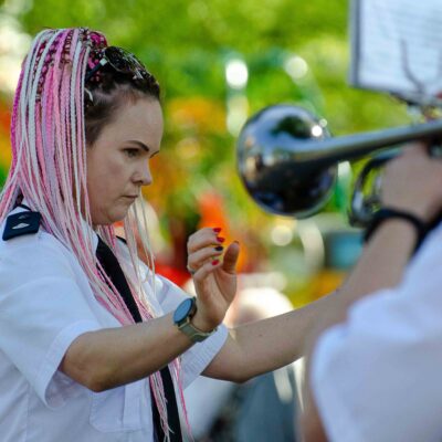 Portret kobiety w białej koszuli mundurowej z różowo-białymi warkoczykami. Kobieta ma skupioną minę i uniesione dłonie, dyryguje. Obok niej widoczny jest fragment srebrnej trąbki i pulpit z nutami.