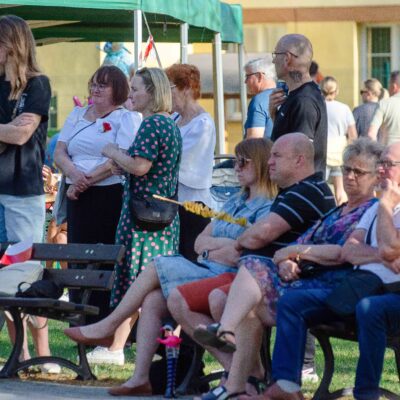 Grupa osób w różnym wieku siedzi na parkowych ławkach i stoi pod zielonym namiotem ogrodowym podczas festynu plenerowego. Ludzie są ubrani w stroje codzienne, panuje swobodna atmosfera.
